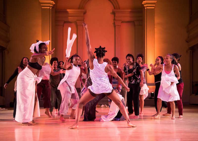 A woman in a white tank top with her back to the audience dances as a group of enthusiastic women cheer her on